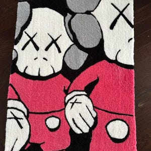 May include: A rectangular tufted rug featuring a graphic illustration of two cartoon-style characters with white skulls, black cross-shaped eyes, and pink clothing. The design includes bold black outlines, white gloved hands, and a gray background.
