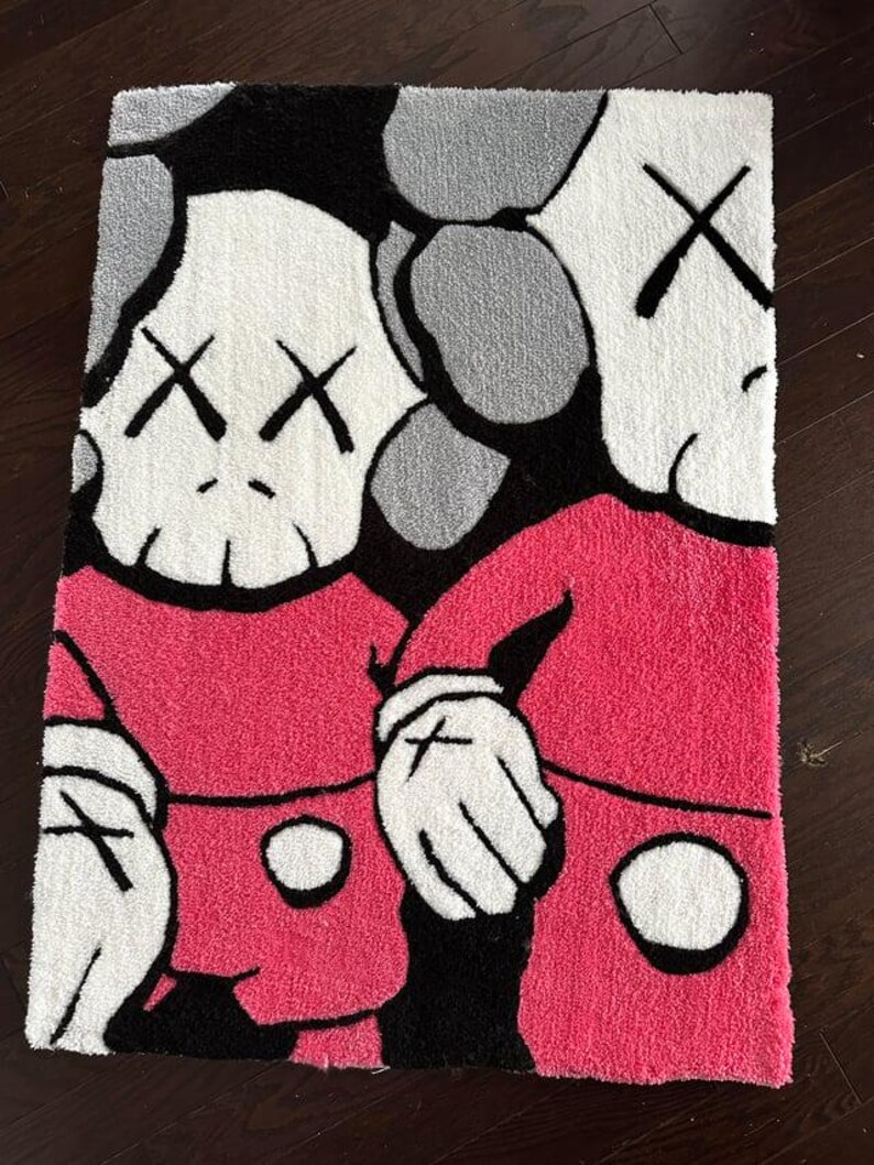 May include: A rectangular tufted rug featuring a graphic illustration of two cartoon-style characters with white skulls, black cross-shaped eyes, and pink clothing. The design includes bold black outlines, white gloved hands, and a gray background.