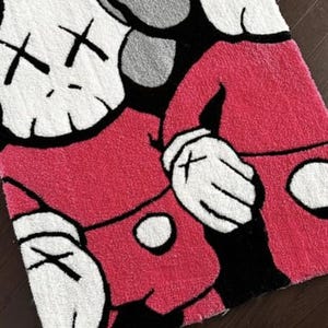 May include: A rectangular tufted rug featuring a graphic design of two cartoon-style figures with white heads, black cross-shaped eyes, and pink bodies. The figures have white gloved hands and are set against a black background with gray accents.