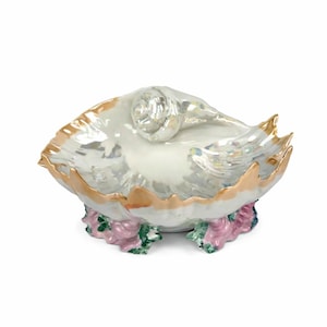 Puede incluir: Un plato decorativo de cerámica con forma de concha y acabado nacarado. El borde está adornado con oro y la base presenta detalles florales rosas y verdes. El plato está diseñado para parecerse a una concha de nautilus.