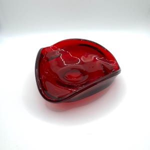 May include: A striking, deep red glass bowl with an unusual, irregular form. The bowl has a smooth, shiny surface and a gently curved edge. The bowl is likely for decorative use or serving small items.