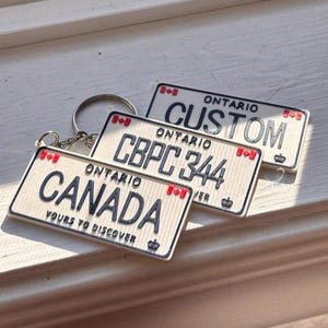 Könnte beinhalten: Drei Schlüsselanhänger mit Nummernschildern aus Ontario. Ein Schlüsselanhänger trägt die Aufschrift "CANADA" mit "YOURS TO DISCOVER" darunter. Ein weiterer trägt die Aufschrift "CBPC 344", und der dritte trägt die Aufschrift "CUSTOM". Jeder Schlüsselanhänger hat eine rot-weiße kanadische Flagge.
