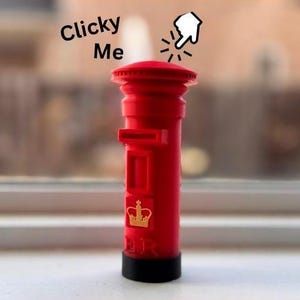 May include: A miniature red British mailbox with a black base. The mailbox has a slot for mail and a gold emblem of a crown. The text "Clicky Me" is above the mailbox.