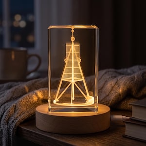 Könnte beinhalten: Ein rechteckiger, klarer Glasblock mit einer internen, per Laser eingravierten 3D-Darstellung eines hohen, spitzen Turms. Der Glasblock steht auf einem runden, hellen Holzsockel, der den Turm in einem warmen gelben Licht erstrahlen lässt.