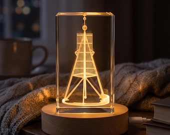 Könnte beinhalten: Ein rechteckiger, klarer Glasblock mit einer internen, per Laser eingravierten 3D-Darstellung eines hohen, spitzen Turms. Der Glasblock steht auf einem runden, hellen Holzsockel, der den Turm in einem warmen gelben Licht erstrahlen lässt.