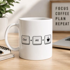 Op de afbeelding: Een witte keramische mok met een afbeelding van drie computertoetsenbordtoetsen op een rij. De toetsen zijn gelabeld met Ctrl, Alt en een icoon van een koffiekopje, gescheiden door plustekens. Het ontwerp is in het zwart op de mok gedrukt.
