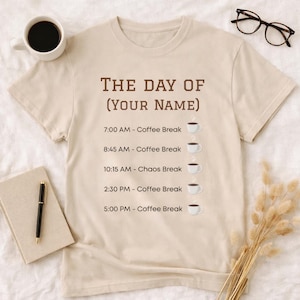 Op de afbeelding: Beige t-shirt met de tekst "THE DAY OF (YOUR NAME)" in bruin. Het shirt toont een schema met koffiepauzes om 7:00, 8:45, 14:30 en 17:00 uur, en een "Chaos Break" om 10:15 uur. Een koffiekopje illustreert elke tijd.