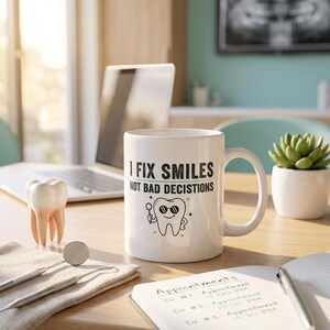 Op de afbeelding: Een witte keramische mok met de zwarte tekst I FIX SMILES NOT BAD DECISIONS en een afbeelding van een lachende tand met een zonnebril die een tandartsspiegeltje vasthoudt. De mok heeft een standaard handvat en een glad, cilindrisch ontwerp.