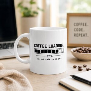 Könnte beinhalten: Ein weißer Keramikbecher mit schwarzem Text, der COFFEE LOADING... lautet, sowie einem Ladebalken bei 75 % und dem zusätzlichen Text Do not talk to me yet. Der Becher hat einen Standardhenkel und ein glattes, zylindrisches Design.