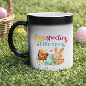 Op de afbeelding: Witte keramische mok met een zwarte handgreep met de tekst "Egg-specting a little bunny" en een illustratie van een konijn, paaseieren en een chocolade-ei. De mok staat op een grasachtig oppervlak met paaseieren en een mand op de achtergrond.