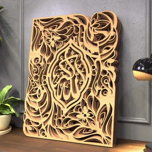 May include: A layered wooden art piece with intricate floral and calligraphic designs. The artwork is made of light brown wood and features a three-dimensional effect. The piece is leaning against a gray wall, with a plant and lamp in the background.