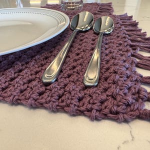 May include: A close-up shot of a lavender crocheted placemat with fringe, a white plate with a gray rim, and two silver spoons. The placemat is made of thick yarn and has a textured appearance. The spoons are placed on the placemat.