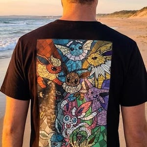 May include: Black t-shirt featuring a colorful stained-glass design of various Pokémon characters. The design includes Eevee and its evolutions, with vibrant colors and intricate details. The shirt is worn by a person on a beach.