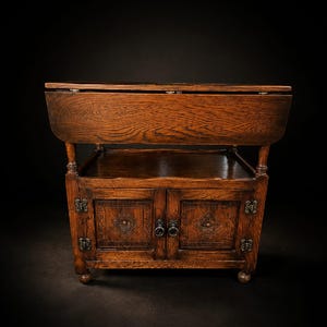 Puede incluir: Una mesa de alas abatibles de madera marrón oscuro con un armario inferior de dos puertas. Las puertas del armario están decoradas con patrones de diamantes tallados y tiradores de anillo metálicos. Se apoya sobre pequeñas patas.