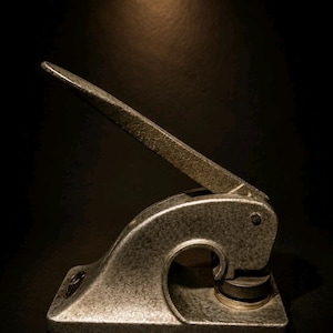 Könnte beinhalten: Ein silberfarbener Schreibtisch-Prägeapparat mit Hebelgriff. Der Prägeapparat hat eine abgerundete Basis und eine kreisförmige Matrize. Das Bild ist vor einem dunklen Hintergrund platziert, der die metallische Textur und das Design des Werkzeugs hervorhebt.