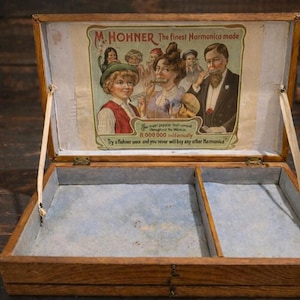 May include: Open wooden box with a hinged lid, featuring an advertisement for M. Hohner harmonicas. The box has two compartments with a light blue lining. The lid displays an illustration of people playing harmonicas. The text reads "The finest Harmonica made."