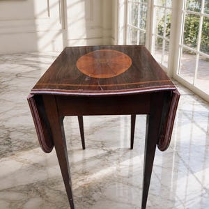 May include: A dark brown wooden drop-leaf table featuring a rectangular top with rounded edges and a central oval inlay with a sunburst pattern. The table stands on four tapered legs and displays a polished finish with visible wood grain details.