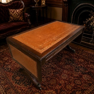 Puede incluir: Un escritorio rectangular de madera con un marco marrón oscuro y una incrustación de cuero color canela en la superficie superior. La incrustación de cuero está decorada con un patrón de borde dorado en relieve. El escritorio tiene dos patas talladas.