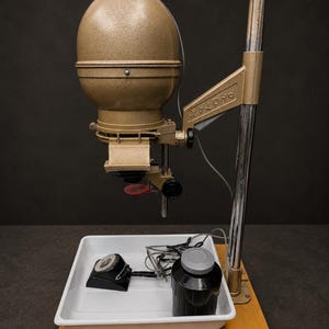 May include: A vintage Photax photographic enlarger with a gold-toned metal housing, mounted on a vertical chrome column and a wooden base. Included are a white plastic developing tray, a black film developing tank, and a small black timer unit.