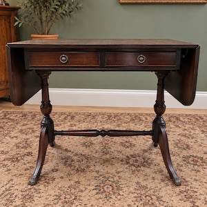 Puede incluir: Una mesa de alas abatibles de madera marrón oscuro con dos cajones horizontales y tiradores metálicos circulares. La mesa se apoya en dos patas de pedestal torneadas unidas por un travesaño central, con pies curvos que terminan en detalles metálicos de garra.