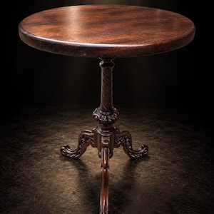 May include: A round, dark brown wooden pedestal table featuring a smooth circular top supported by a central column with a twisted design. The base consists of three ornate, curved legs with intricate carved detailing at the junction point.