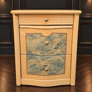 Puede incluir: Una mesita de noche de color crema claro con un cajón superior de ranuras horizontales y dos cajones inferiores con incrustaciones de patrón de mármol azul y crema. Incluye pequeños pomos redondos de metal en tono dorado.