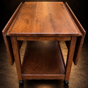 Puede incluir: Una mesa auxiliar rectangular de madera marrón con diseño de alas abatibles en ambos lados, un estante de almacenamiento inferior y cuatro pequeñas ruedas negras en la base. La veta de la madera es visible en la superficie superior.