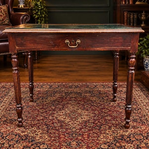 May include: A dark brown wooden writing desk featuring a rectangular top with a green leather inlay surface. The desk includes a single central drawer equipped with a brass pull handle and a keyhole. It stands on four turned, fluted legs.