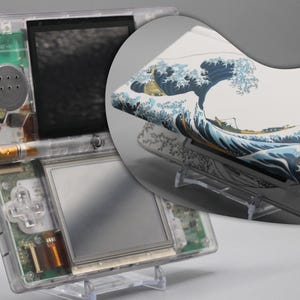 Könnte beinhalten: Eine tragbare Spielkonsole mit einem transparenten Kunststoffgehäuse, das interne Komponenten wie einen Lautsprecher und eine Platine zeigt. Die Außenseite ist mit einem Druck einer großen blauen Welle mit weißem Schaum versehen.
