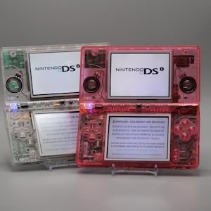 Peut inclure: Deux consoles de jeux portables Nintendo DSi avec des coques transparentes, l'une incolore et l'autre rose translucide. Les deux appareils disposent de deux écrans, les écrans supérieurs affichant le logo Nintendo DSi et les inférieurs du texte.