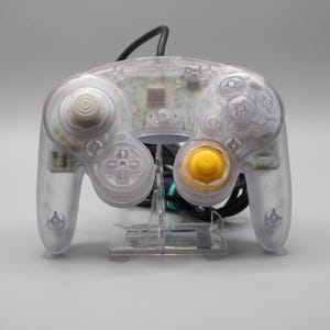 Può includere: Un controller per videogiochi trasparente e incolore dotato di una levetta analogica sinistra bianca, una levetta analogica destra gialla, un pad direzionale e vari pulsanti di azione. Il circuito stampato interno è visibile attraverso l'involucro.