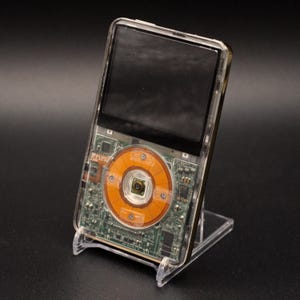 May include: A clear, transparent iPod with its internal components visible, including a circular orange component and green circuit board. The iPod is displayed on a clear plastic stand against a black background.