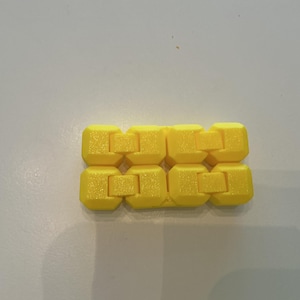 May include: A bright yellow fidget toy composed of eight connected, interlocking blocks. The blocks have a textured surface and are arranged in a rectangular shape. The toy is designed to be manipulated and played with.