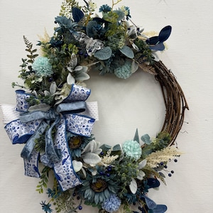 May include: A decorative wreath featuring a variety of blue and green faux flowers and foliage. The wreath includes a large bow with a blue and white floral pattern. The base is made of woven twigs, creating a natural look.