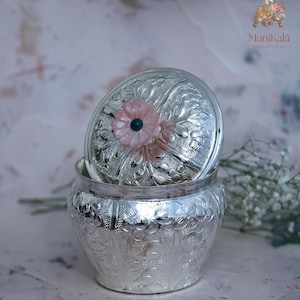 May include: A silver-colored, ornate jar with a lid, featuring a floral design. The lid is slightly ajar, revealing the interior. A pink flower with a green center adorns the lid. The jar is likely used for storing small items. The brand name "Manikala" is visible.