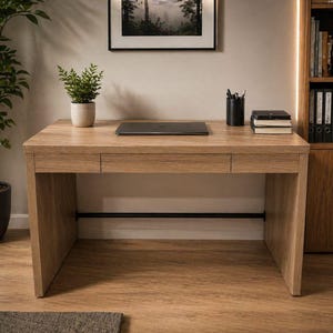 May include: A light brown wooden desk with a closed laptop, a small potted plant, and a black pen holder. The desk has a drawer and a black metal bar for support. A framed picture hangs on the wall above the desk.