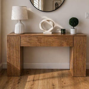 May include: A wooden console table with a ribbed design, topped with a white lamp, a white sculpture, and a small potted plant. The table is set against a neutral wall with a round mirror and a window.