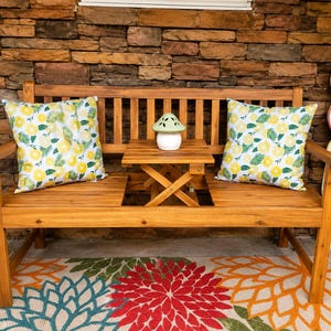 May include: A wooden bench with a built-in center table, featuring two square pillows with a yellow and green citrus fruit pattern. A small, light green mushroom-shaped ceramic decor piece sits on the table. A vertical sign reads WELCOME.