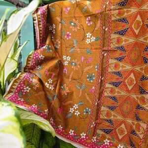 May include: A mustard yellow fabric featuring a floral pattern with white, pink, and teal blossoms on thin vines. The fabric includes a vertical border with a geometric diamond design in orange, dark blue, and white, finished with a pink floral trim.