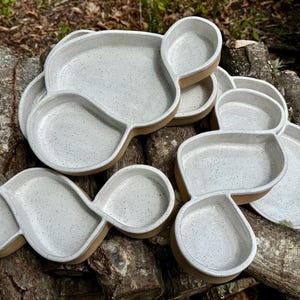 May include: A collection of handmade ceramic serving platters. Each platter features a unique, organic shape with multiple compartments. The platters have a speckled, light gray interior and a warm, brown rim. Perfect for serving appetizers or snacks.