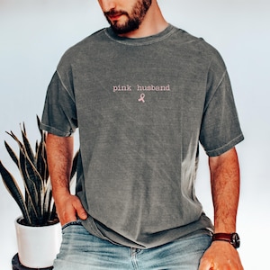 May include: A gray t-shirt with the text "pink husband" in pink lettering and a pink ribbon graphic. The shirt has short sleeves and a relaxed fit. The model is wearing blue jeans and a brown watch.
