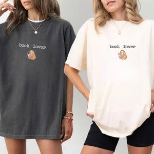 Puede incluir: Dos camisetas holgadas, una gris antracita y otra crema, con el texto "book lover" sobre un pequeño gráfico de un libro en una cesta. Las camisetas tienen mangas cortas y un corte relajado.