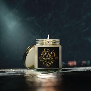 Puede incluir: Una vela encendida en un tarro de cristal con tapa dorada. La etiqueta negra presenta el texto dorado "Eid Mubarak" con elementos decorativos. La vela se encuentra sobre un fondo oscuro y misterioso.
