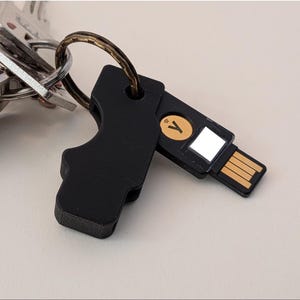 May include: A black security key with a gold-colored USB connector and a circular gold logo featuring a Y symbol and a wireless icon. The device is attached to a metal keyring along with a collection of silver-colored metal keys.