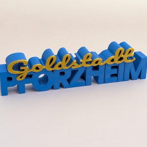 May include: A three-dimensional sign featuring the word PFORZHEIM in bold blue capital letters, with the word Goldstadt written in a gold cursive script layered on top of the blue text, creating a layered and raised decorative display piece.