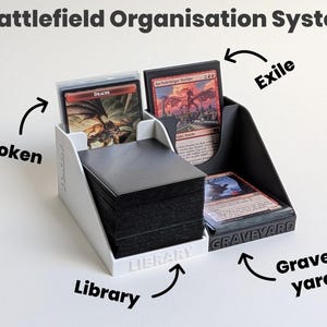 Könnte beinhalten: Ein Battlefield Organisation System Kartenhalter mit einem weißen Bereich beschriftet mit Library und Token, sowie einem schwarzen Bereich beschriftet mit Exile und Graveyard. Der Halter enthält Stapel von Spielkarten in schwarzen Hüllen.