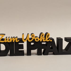 Op de afbeelding: Een zwart en geel 3D-bord met de tekst "Zum Wohl. DIE PFALZ". De letters "Zum Wohl" zijn geel, de rest is zwart. Dit decoratieve bord is waarschijnlijk bedoeld om te worden tentoongesteld.