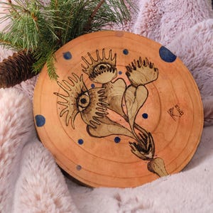 May include: A round, wooden art piece with a burnt sienna finish. The artwork features a stylized floral design with an eye and other organic shapes. Dark blue dots are scattered around the design. A pine branch and cone are in the background.