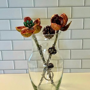 May include: A clear glass vase containing decorative floral stems. The stems feature orange and brown dried flowers and small pinecones. The stems have a glittery, silver coating. The vase is set against a white tiled background.