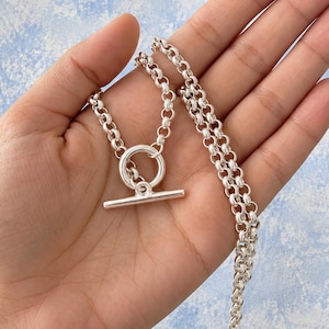 May include: A silver-toned necklace featuring a thick rolo chain and a toggle clasp closure. The necklace consists of a circular ring and a horizontal bar component that secures the chain, showcasing a polished metallic finish throughout the piece.